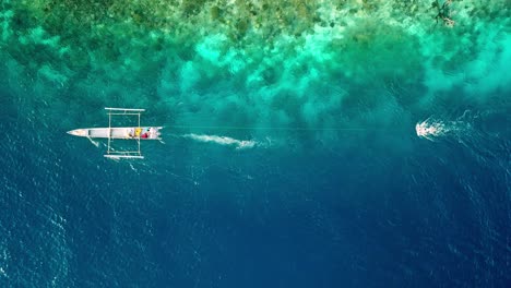 Diver-ocean-gliding-with-a-Subwing-in-Raja-Ampat-Indonesia-coral-reef-island-pulled-by-a-small-motorboat,-Aerial-top-view-tracking-left-lowering-shot