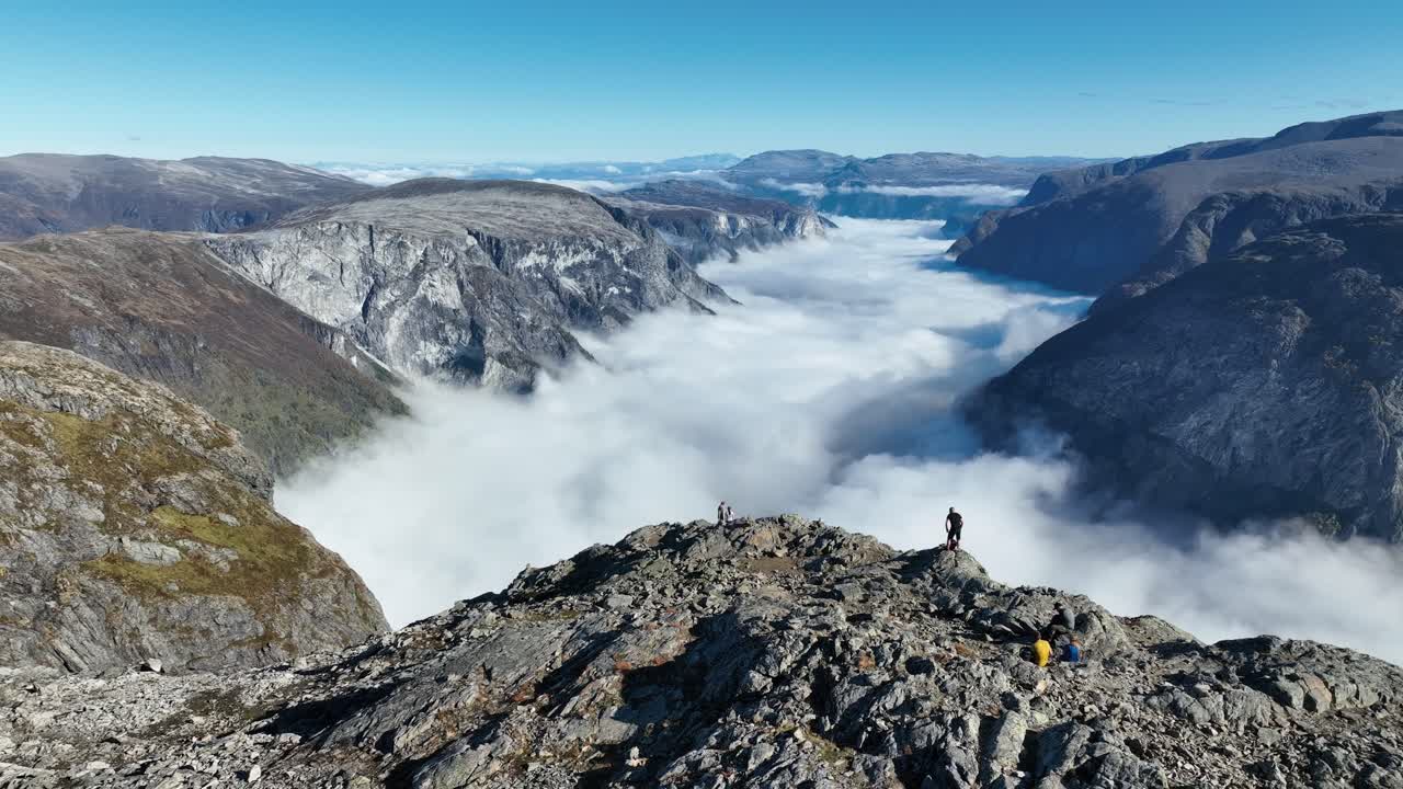 Drone orbits from above, showing a couple and other tourists at the cliff edge of Bakkanosi with stunning views of the fjord and fog below