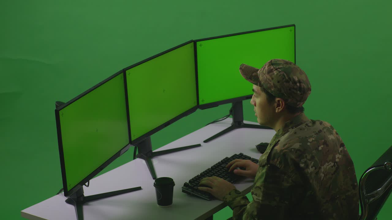 vista lateral de un soldado asiático cansado bostezando mientras trabaja con un simulacro de múltiples monitores de computadora en un estudio de pantalla verde