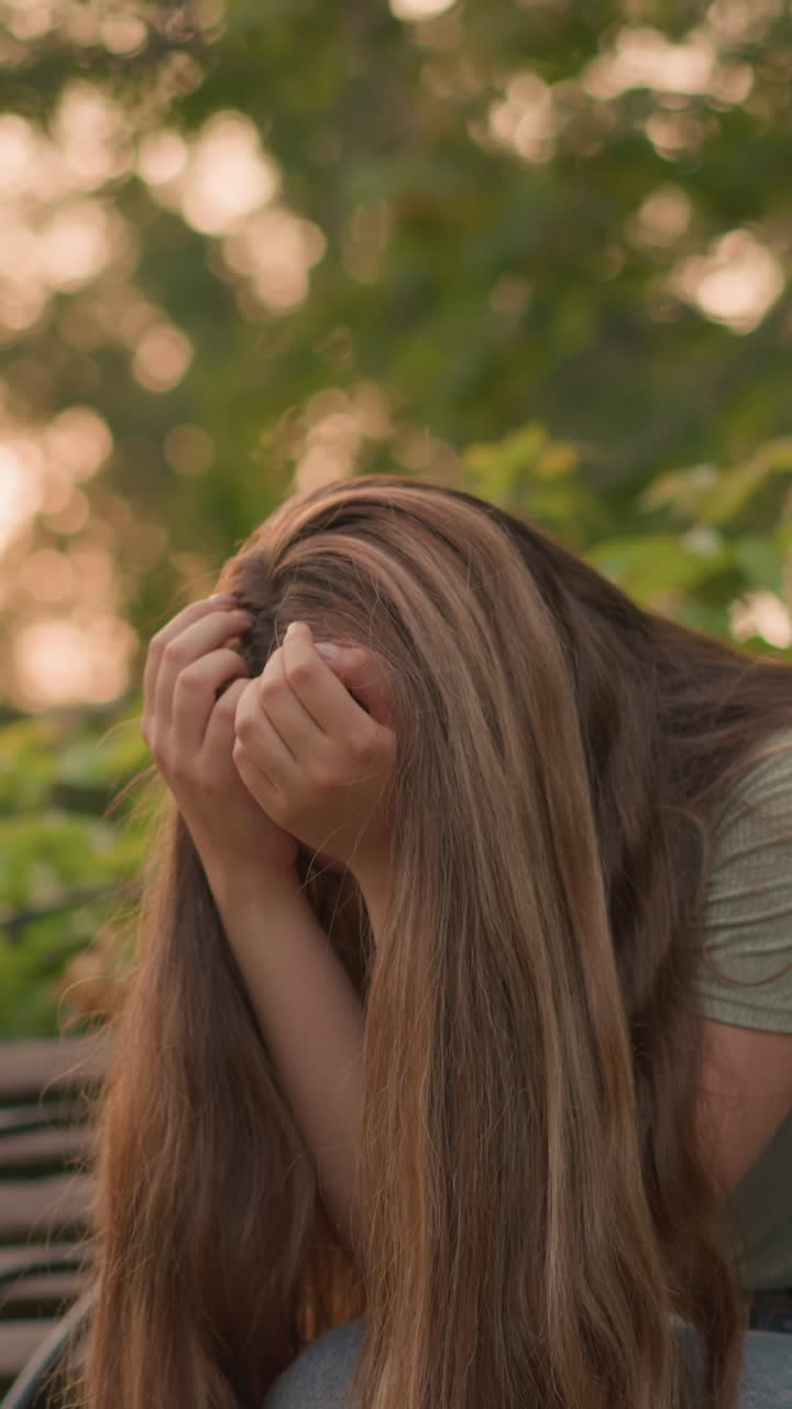 mujer sentada en un banco de madera en una postura reflexiva y sombría, manos cubriendo la cara, su largo cabello cayendo en cascada por los hombros, la cálida luz del sol filtrándose a través de los árboles y la exuberante vegetación