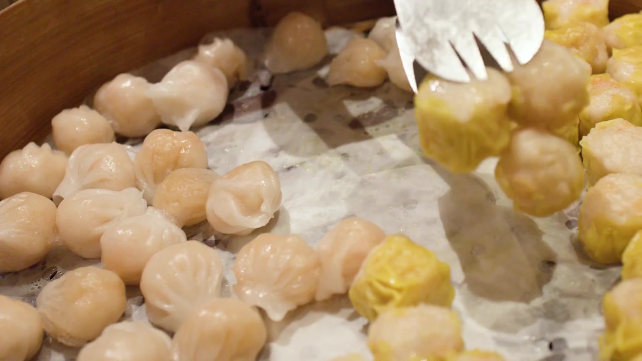 Potret jarak dekat penjepit logam memilih pangsit shumai kuning dari keranjang kukusan bambu besar