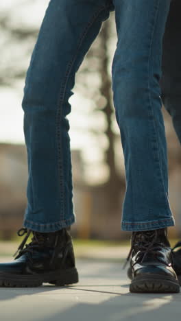 un hombre está dejando caer a una niña pequeña. la niña lleva pantalones vaqueros azules y botas negras, y las piernas del hombre son visibles en el marco. están al aire libre, con árboles y luz solar en el fondo