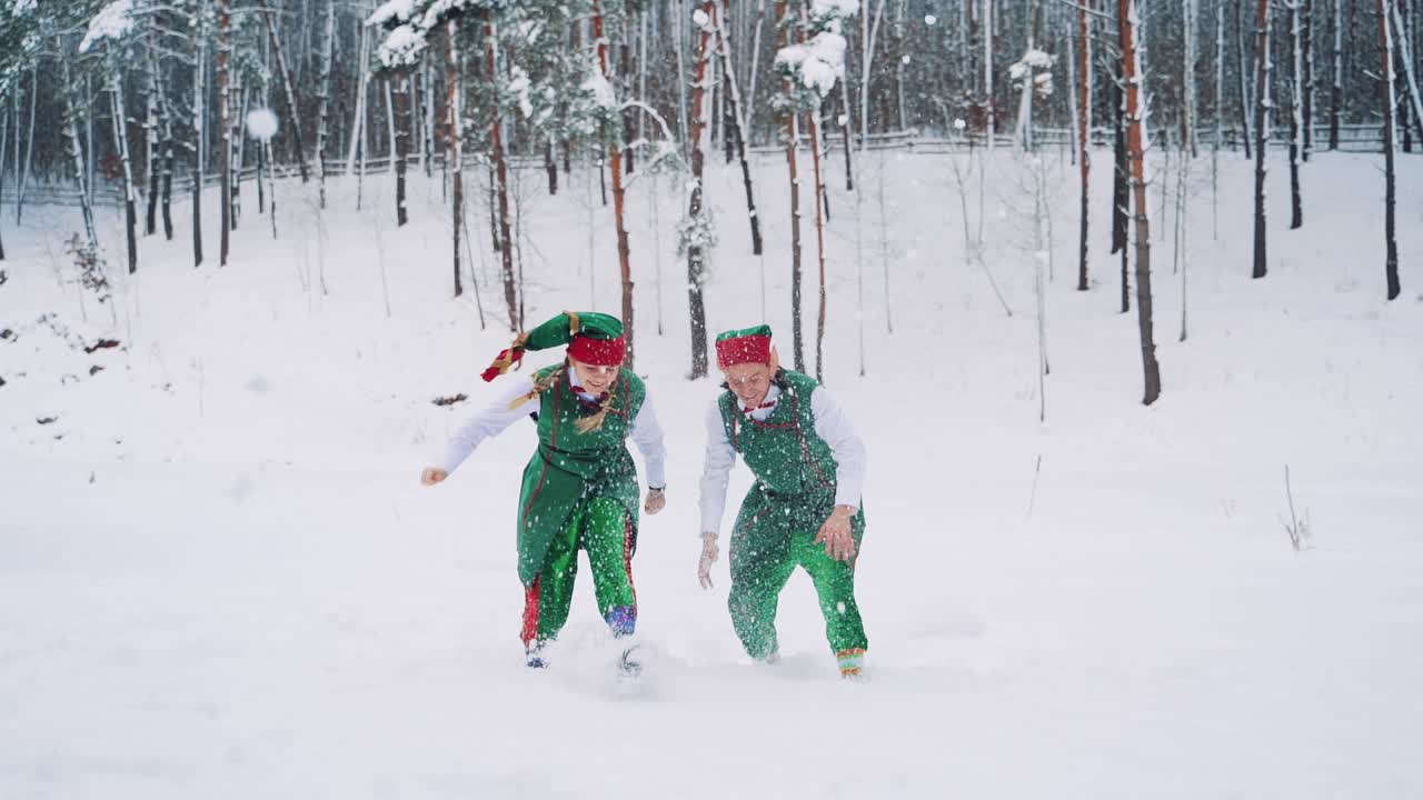 Two elves in green suits with hats are playing and throwing up snow with their hands on the background of the winter forest. Slow motion