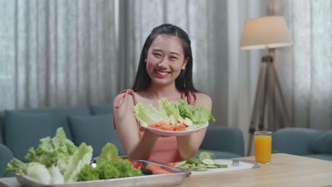 smiling asian woman showing a dish of healthy food to camera while sitting at home