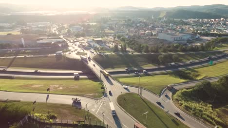 Luftdrohnenansicht-Von-Großen-Straßen-Und-Kreuzungen-Mit-Viel-Verkehr-Inmitten-Von-Grünflächen-Und-Einer-Kleinen-Stadt-Und-Bergen-Im-Hintergrund