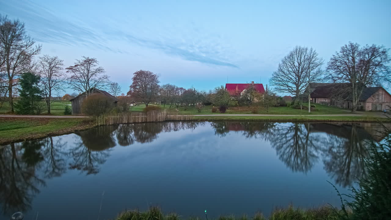 Timelapse af solnedgang ved sø på en grøn eng med træer og træhuse og skyer, der trækker ind i horisonten.