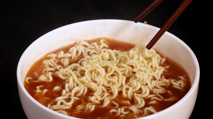 Bacchette sollevano noodles istantanei fumanti da una ciotola di brodo su uno sfondo scuro da studio.