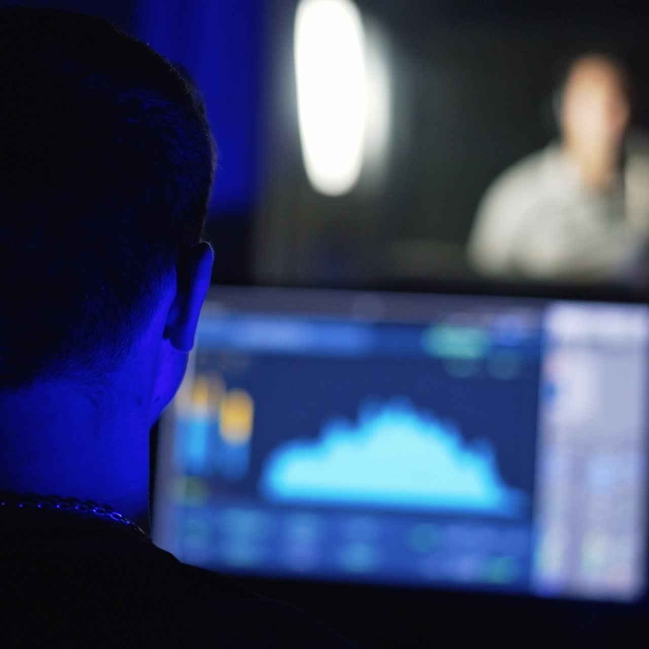 Sound engineer's rear view sitting in the recording room in front of equipment. A pair of people behind the glass performing song. Blurred backdrop