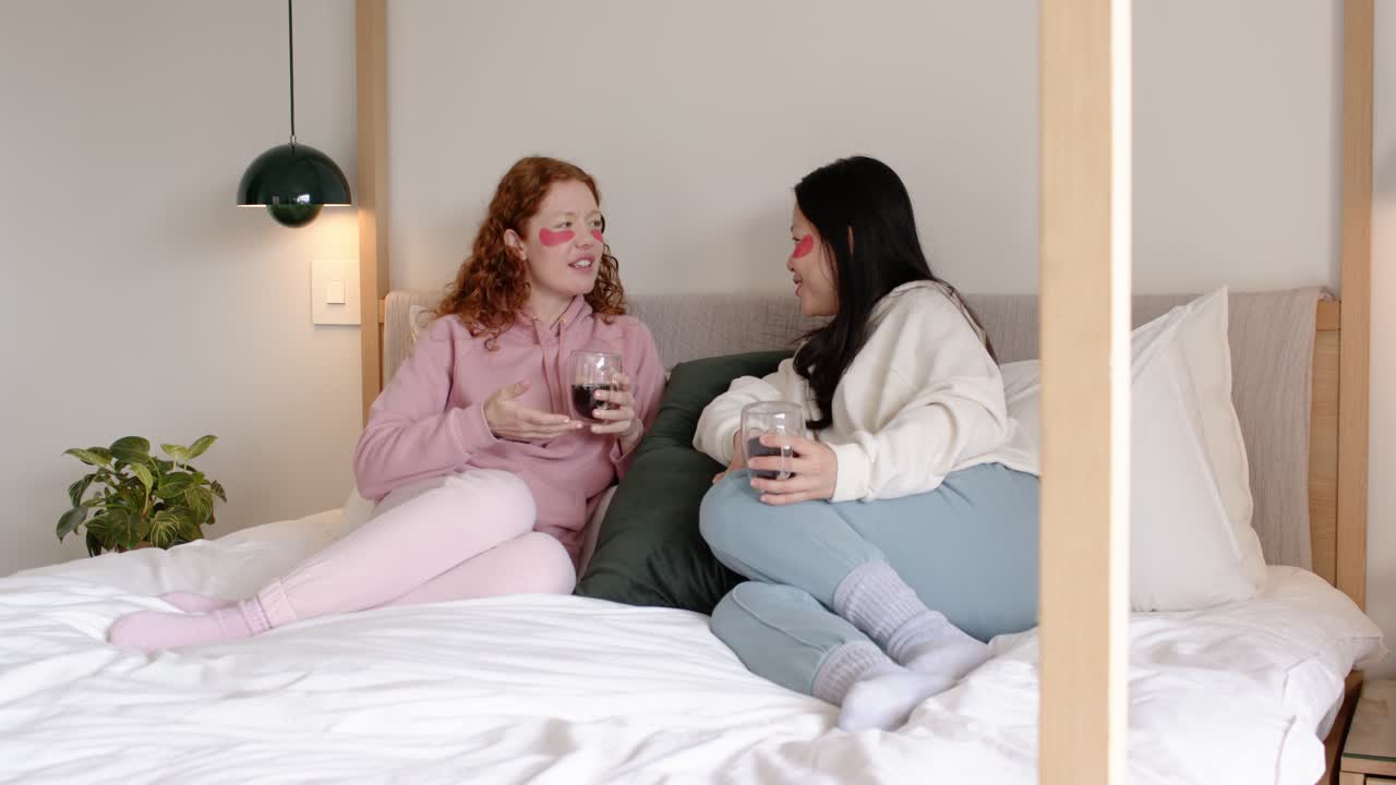 Diverse female friends sitting on canopy bed, left woman leading chat gesturing with glass to bond