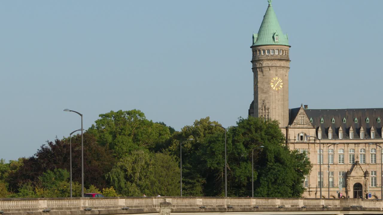 Green tractor drives across scenic stone bridge past historic clock tower, daylight, wide shot