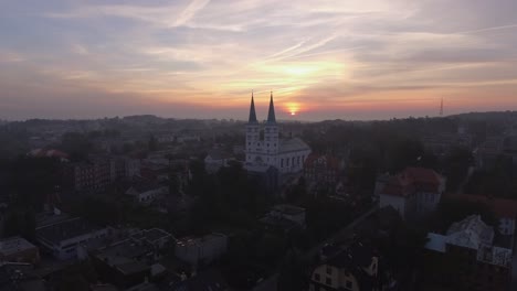 Toma-Aérea-De-Una-Iglesia-Cristiana-En-Una-Pequeña-Ciudad-Europea-Durante-El-Amanecer-Brumoso