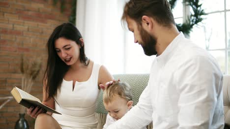 Young-mother-reading-book-for-her-son-and-husband.-Family-reading-Christmas-story-while-sitting-by-Christmas-tree.-Happy-toddler