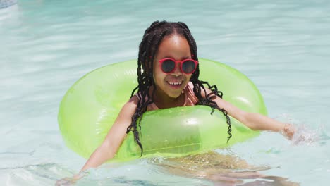 Happy-african-american-girl-wearing-sunglasses-swimming-with-inflatable-ring