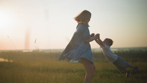 a joyful scene of a mother in a flowing blue gown spinning her young son, who is dressed in a white top and jeans, in a grassy field during sunset