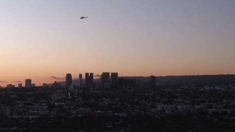 Helicóptero-Sobrevolando-El-Horizonte-Del-Centro-De-Los-Ángeles-Durante-La-Puesta-De-Sol,-Toma-Aérea-Descendente