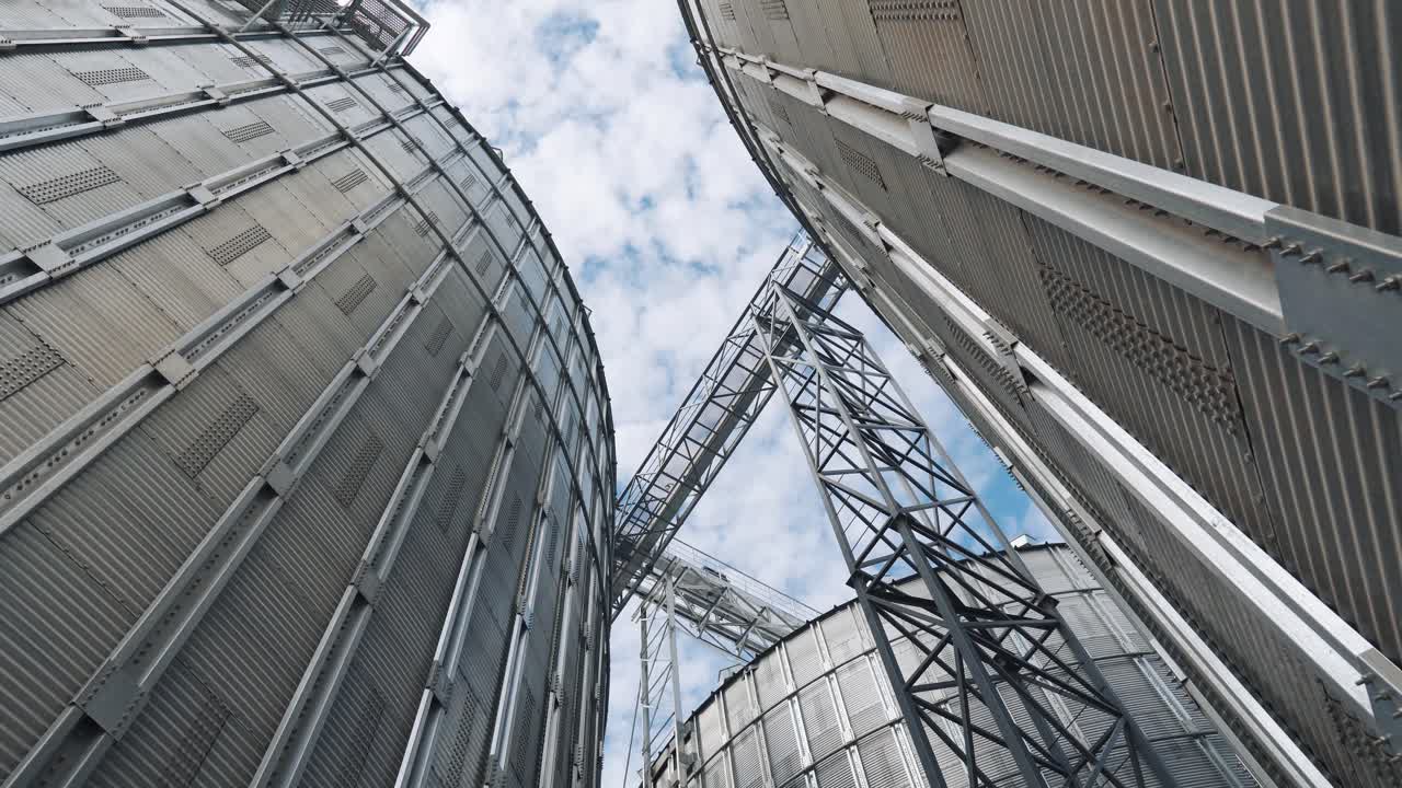 Large grain elevators. Silver granary for storing crop. Metal grain terminals outdoors. Agricultural warehouses. View from below