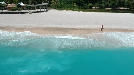 El-Dron-Que-Se-Cierne-Sobre-El-Océano-Ofrece-Una-Vista-De-La-Gente-En-Una-Playa-Lejana-Y-El-Agua-Increíblemente-Azul-Del-Mar-Caribe-Rodando-Hacia-La-Orilla