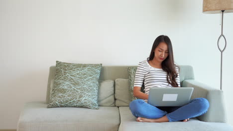 una joven asiática sentada en un elegante sofá sonríe mientras escribe en su laptop