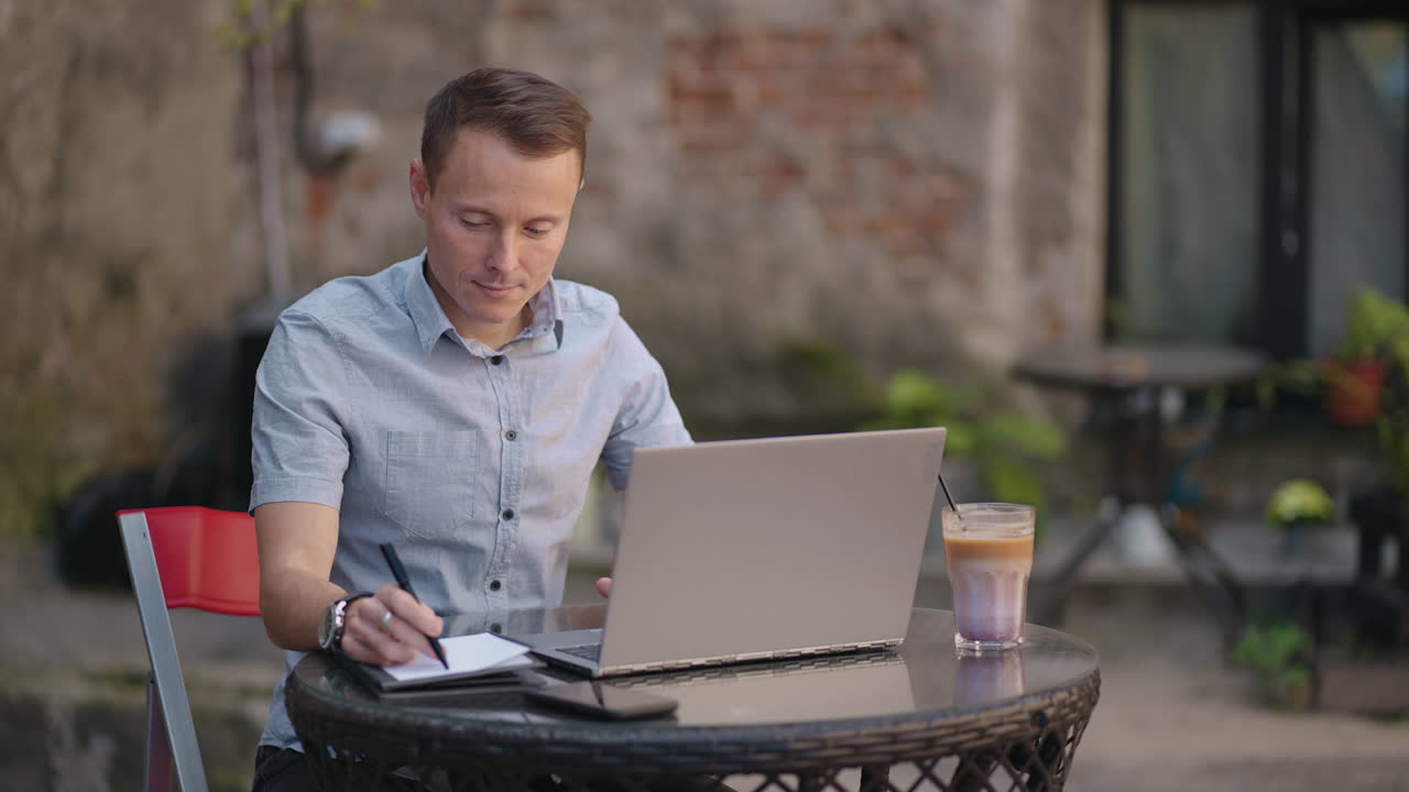 A man sitting in a street cafe works remotely writing a pen in a notebook. Work at the laptop. A man works remotely on a laptop
