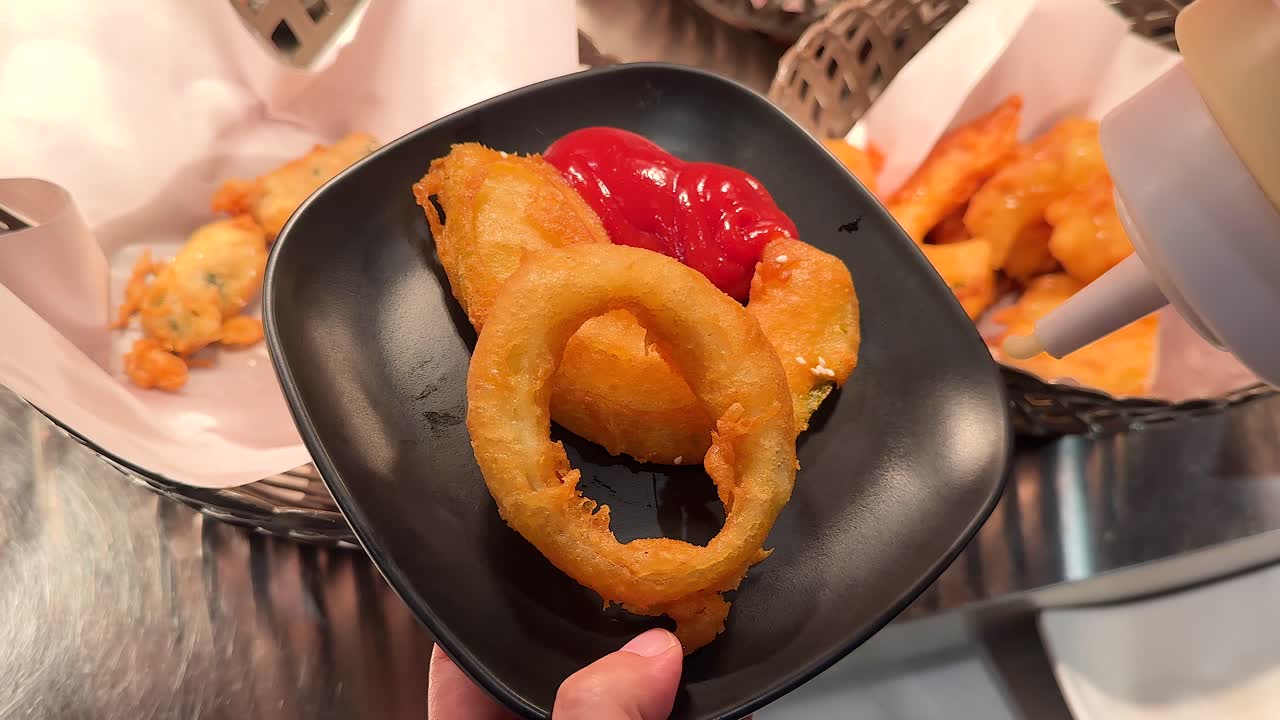 Close-up of fried onion rings on a black plate with ketchup and mayonnaise, set in a vibrant restaurant environment