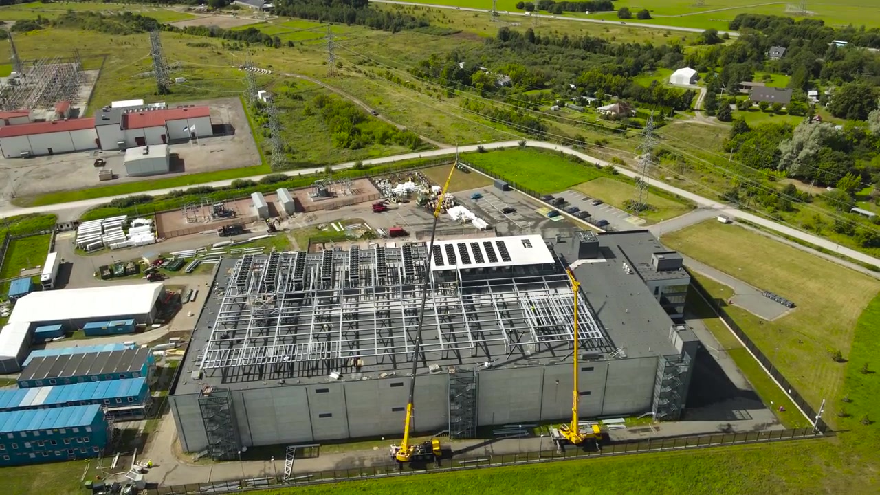 Perspectiva de ângulo alto mostrando guindastes móveis montando a estrutura metálica no telhado de um grande centro de dados, com uma subestação elétrica e uma paisagem verde exuberante ao fundo.