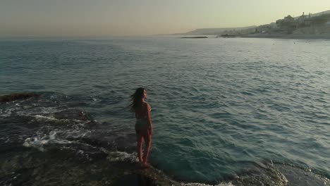 young caucasian woman with long brown hair wearing bathing suit stands on batroun rock outcropping island in blue ocean sea water at sunset, lebanon, overhead aerial flyover approach