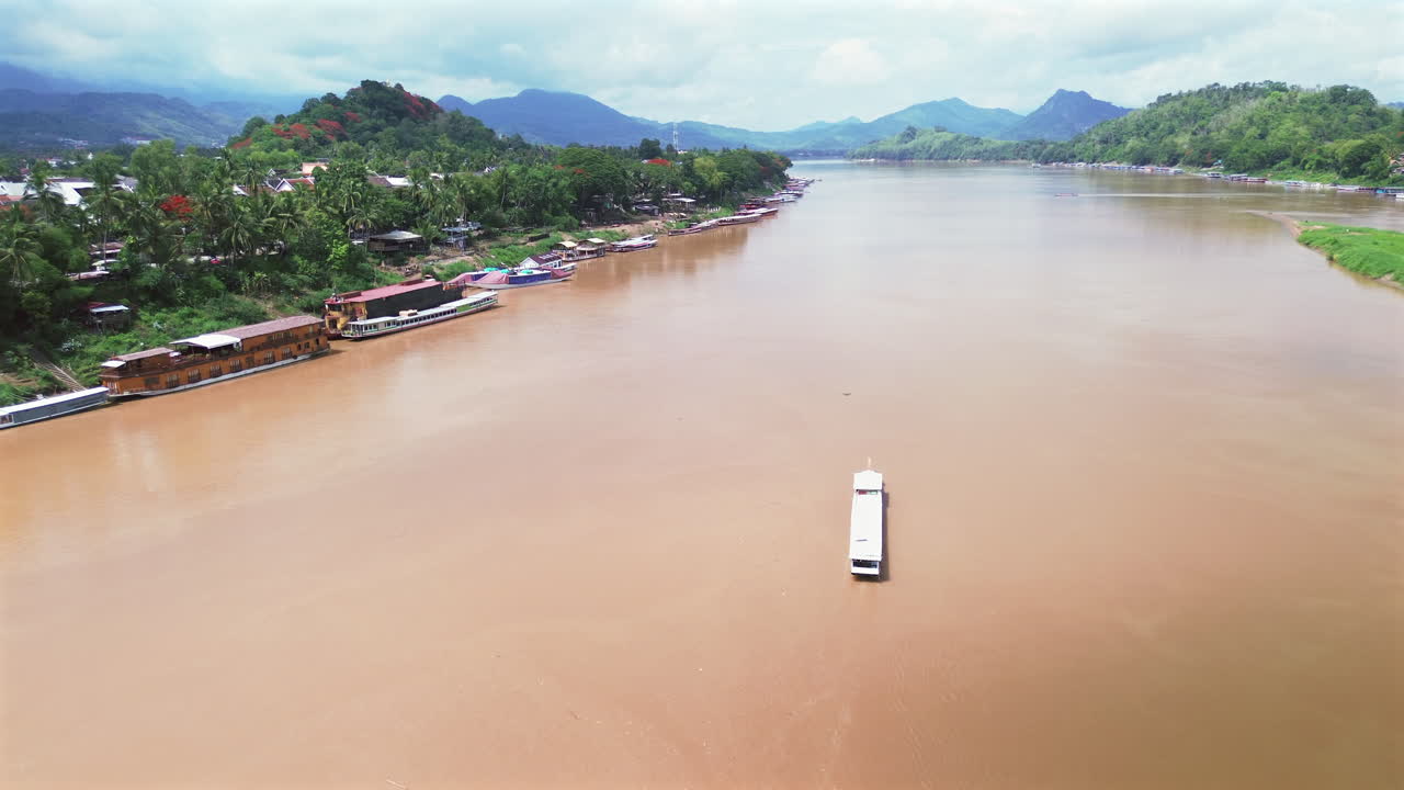 Long wooden boat moves slowly on Mekong River toward Luang Prabang, Laos, under soft light, aerial overview
