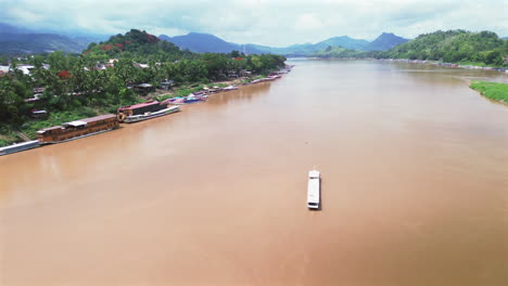 long wooden boat moves slowly on mekong river toward luang prabang, laos, under soft light, aerial overview