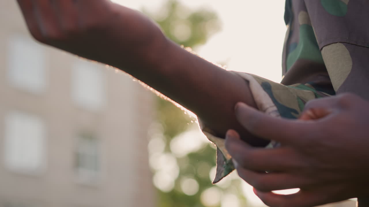 Youthful Military Pride, Cadet Preparing Uniform At Dusk, Hand Securing Wristband During Evening Training, Cadet Tightening Armband On Camouflage Attire During Sunset On Grassy Open Area