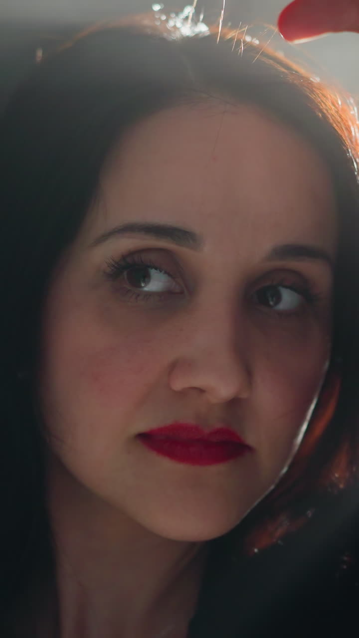 intenso primer plano del rostro de una mujer con llamativos labios rojos, enmarcado contra un fondo oscuro y suavemente iluminado, capturando un momento de profunda expresión