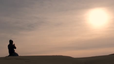 a man kneels to pray to the heavens, while alone in the desert under a beautiful pink sunset