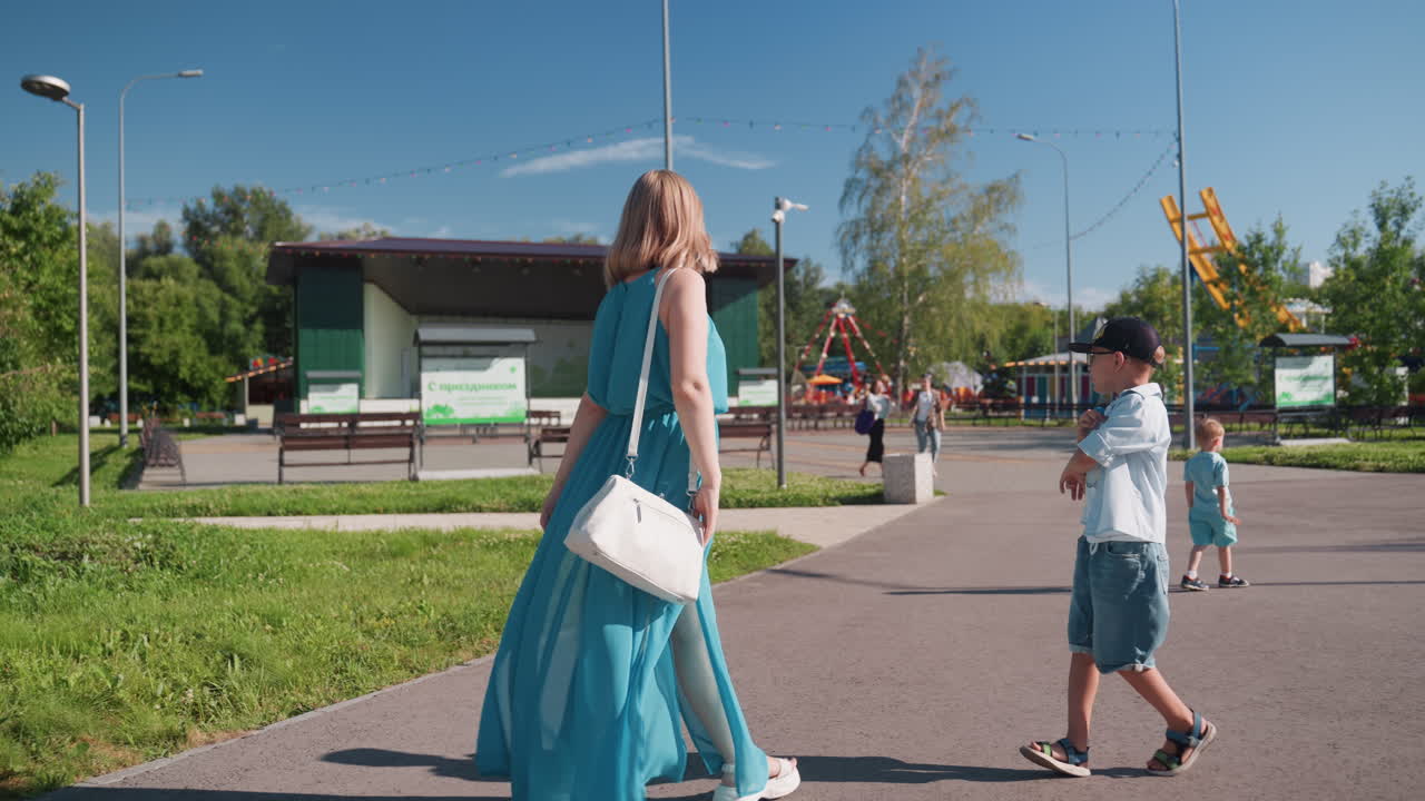 Mutter geht mit Kindern im Park, trägt ein fließendes blaues Kleid und eine Handtasche, ein Junge mit Kappe folgt, ein Kleinkind wandert auf einem grasbewachsenen Pfad, sonniger Wochenend-Familienausflug, stilvolle, legere Stimmung, sanfte Brise, authentische Gehaufnahmen.