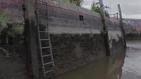 drone view over dark water of old harbor with shabby brick walls and metal ladders in modern city