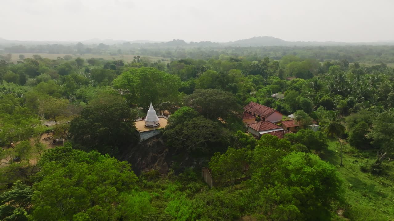 Sri Lanka aerial reveal of a white stupa set on a rock outcrop above dense green canopy with soft morning haze over surrounding countryside