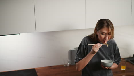 young woman in casual gray shirt eating noodles with chopstick from bowl, bending slightly forward with focused look, glass of orange juice beside her on wooden kitchen counter