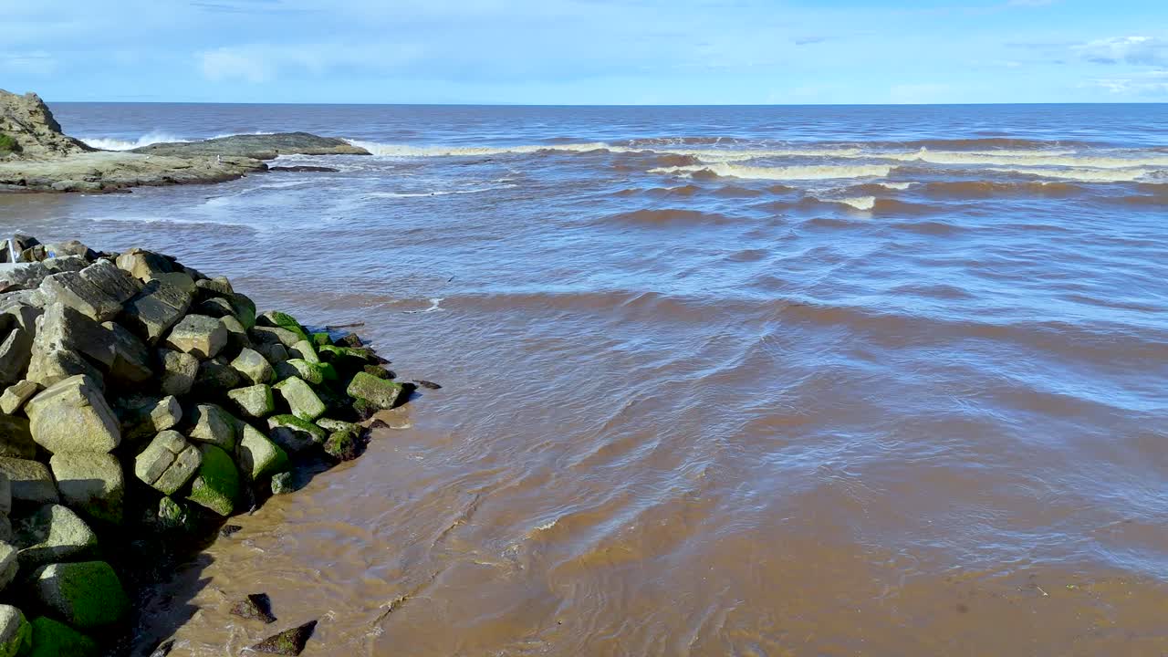 Aerial drone view of stone breakwater where brown river meets ocean waves in bright daylight