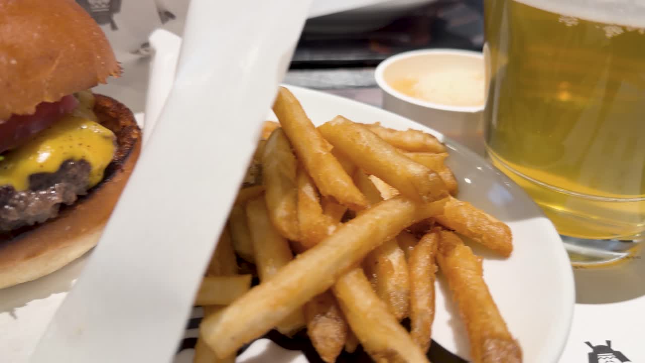 Movimento de câmera na mão em close-up explora um cheeseburger gourmet, batatas fritas crocantes e cerveja lager gelada.