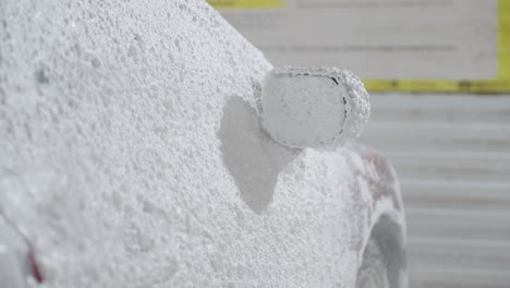 side close up of soapy foam splashing and covering side mirror of red car during washing process with high pressure spray forming thick white bubbles across vehicle surface inside garage environment