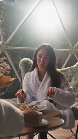 Woman listens to man eating pancakes. Couple discusses day schedule at breakfast time in glamping residence with view on nature. Tranquil morning together