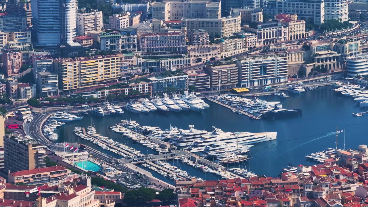 Il porto di Monaco mostra un vivace porto turistico pieno di yacht. Gli edifici circostanti evidenziano l'architettura lungomare della città. Durante il giorno è visibile il passaggio di persone lungo le banchine.