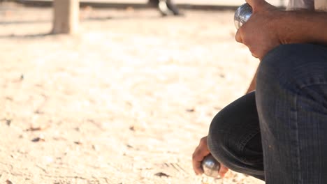 primer plano de un jugador de petanca sosteniendo 2 bolas de metal en la mano y disparando durante un juego de petanque, en un suelo de tierra en un parque soleado en parís en la tarde de verano.