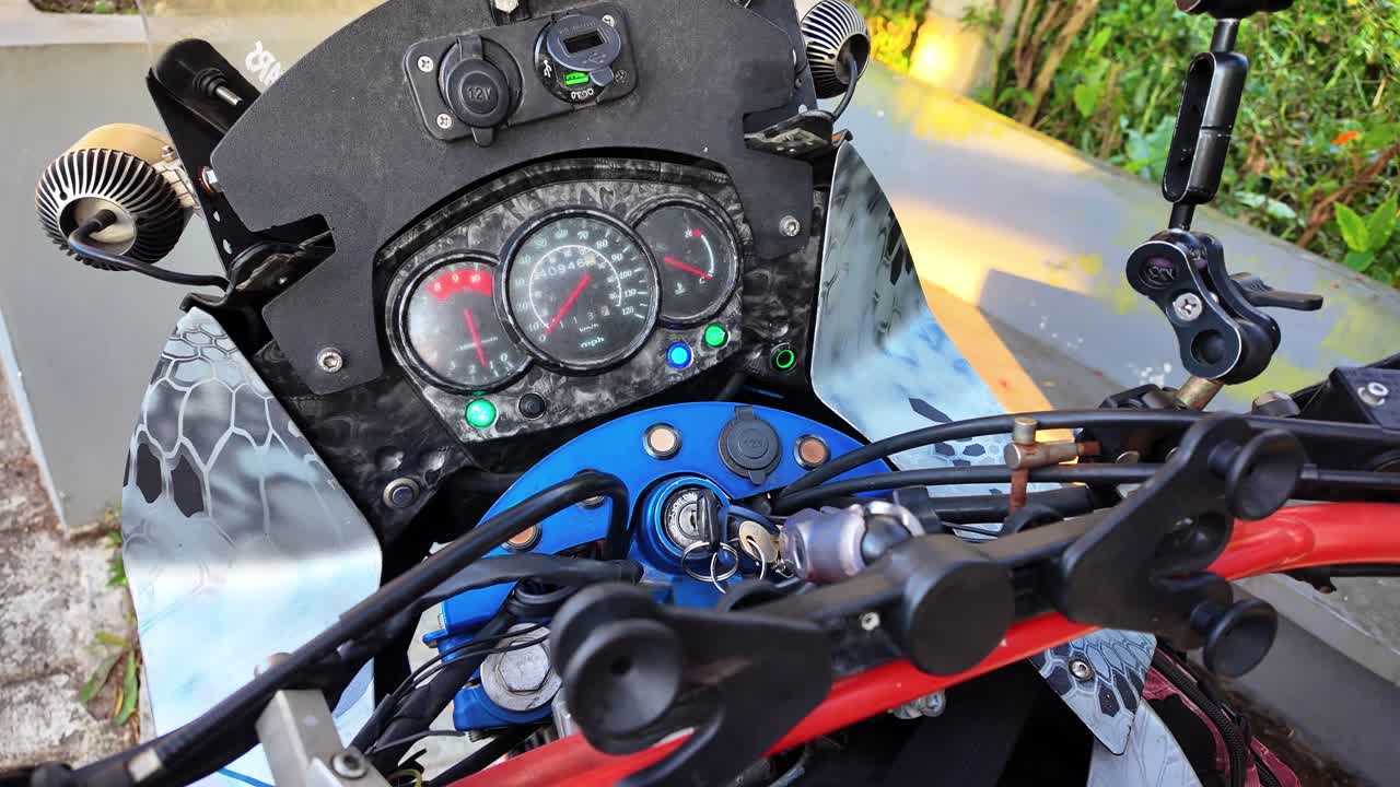 Inquadratura cinematografica in soggettiva del cruscotto di una moto dual-sport che si illumina mentre il motociclista si prepara per un viaggio all'aperto attraverso paesaggi montani.