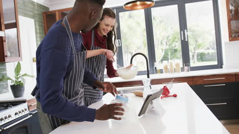 Feliz-Pareja-Diversa-Parada-En-La-Cocina,-Usando-Tableta-Y-Preparando-Masa,-Cámara-Lenta