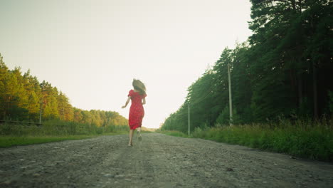 rear view of woman in red floral dress running on unkept dirt road through forested countryside, with hair flowing and sunlight casting warm glow on surrounding trees under wide open sky