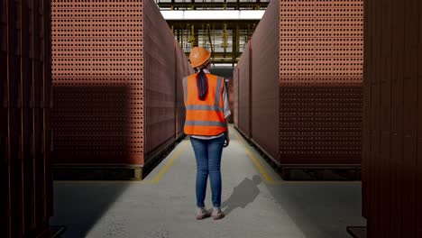 full body back view of asian female engineer with safety helmet talking on smartphone while standing with red brick packed in stacks are stored