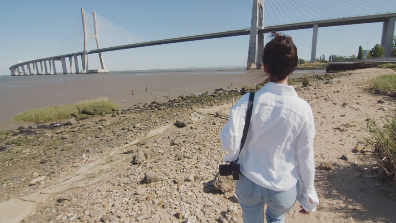 Follow shot of a young woman walking by a small rocky reach near Tagus river and Vasco da Gama bridge in Lisbon, Portugal