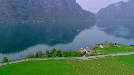 Aerial-view-of-idyllic-shoreline-with-path,-cruising-motorboat-on-Fjord-and-mountain-range-in-background-in-Norway