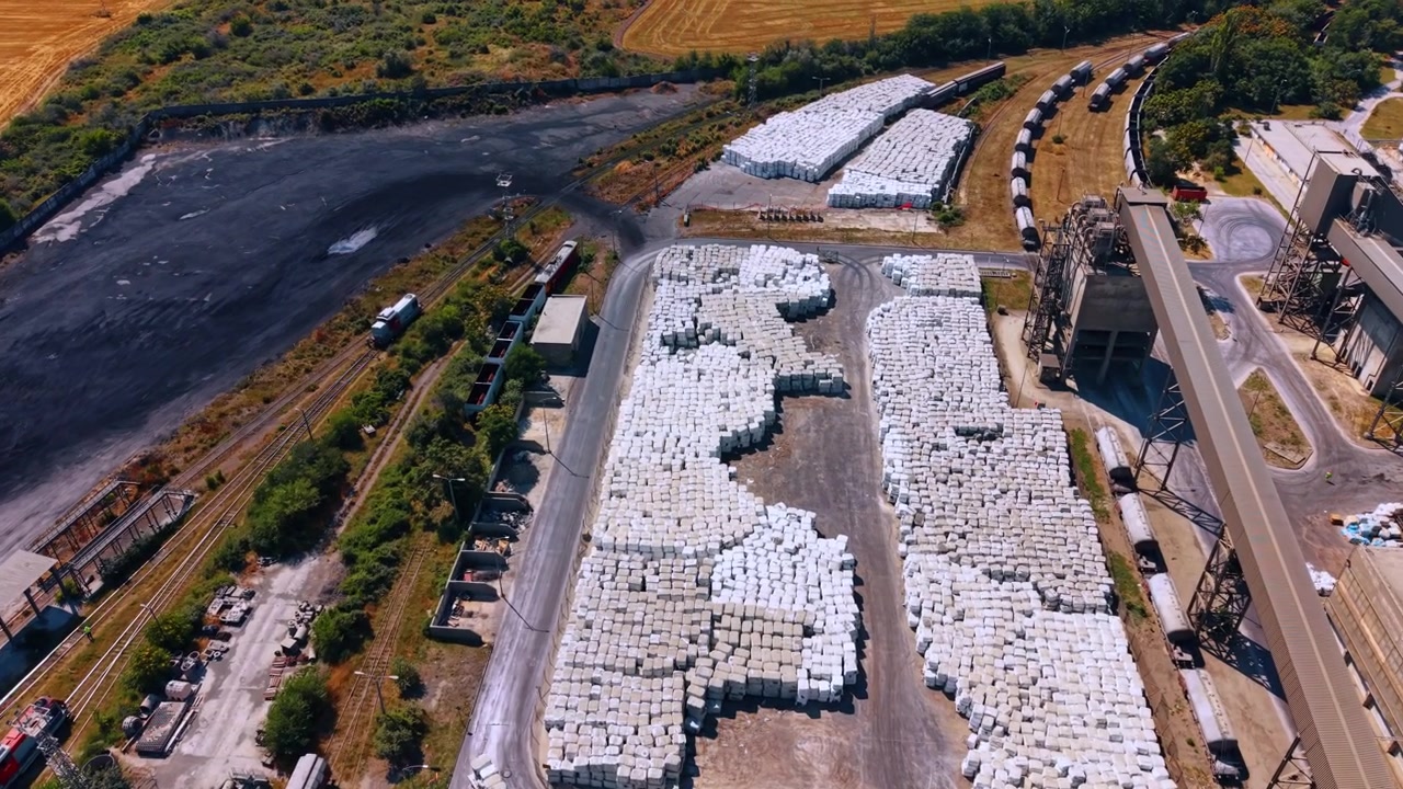 Wrapped materials stored beside industrial rail yard. Large stacks of white packages fill a paved storage area beside freight cars and enclosed conveyors.