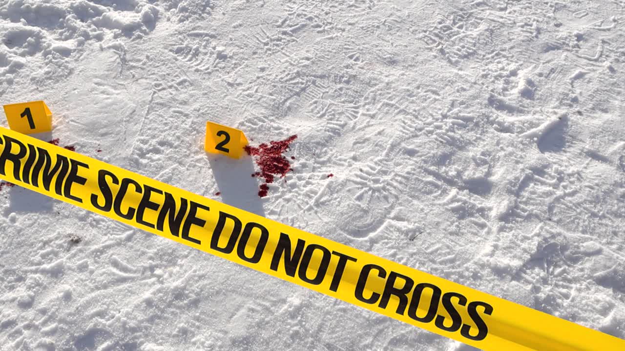 Overhead glide above police crime scene tape and numbered evidence markers beside scattered blood stains on sunlit snow during winter investigation. Sunlight highlights the textured snow covered road