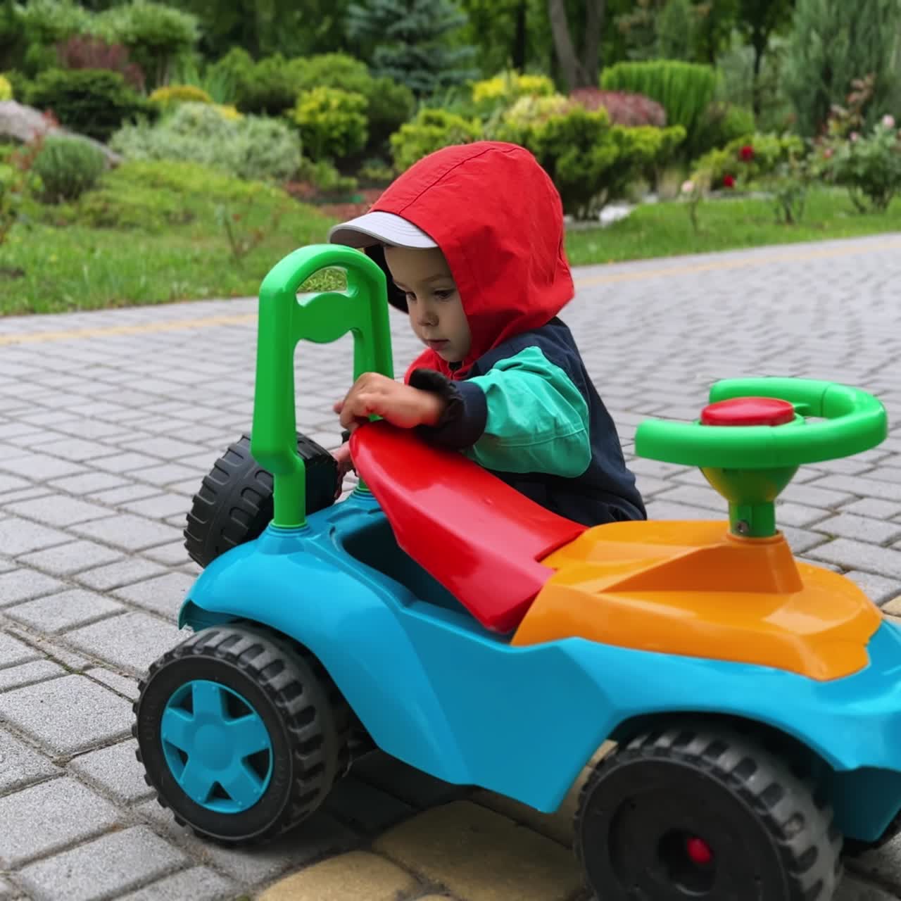 Bambino adorabile e carino con giacca e cappuccio in piedi vicino alla sua macchinina. Il bambino tocca le parti dell'auto nel parco